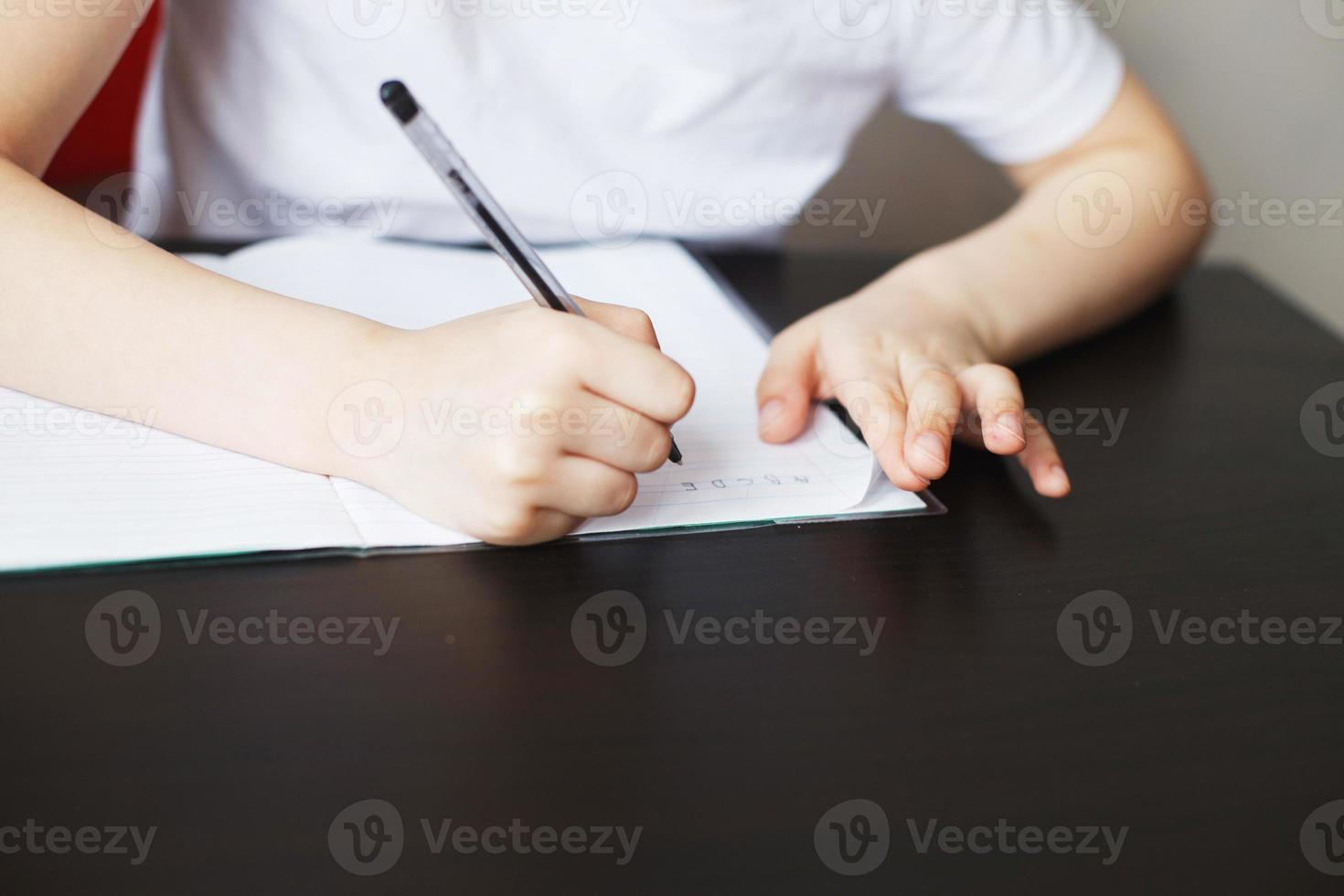 the boy sits at the table and writes in a notebook. child sits and does homework on a white background photo
