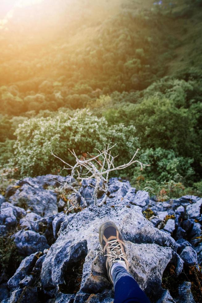 cerrado arriba excursionismo Zapatos de mujer y en pie en rock acantilado mientras hermosa amanecer brillante a bosque en el montaña. éxito vida y libertad concepto. foto