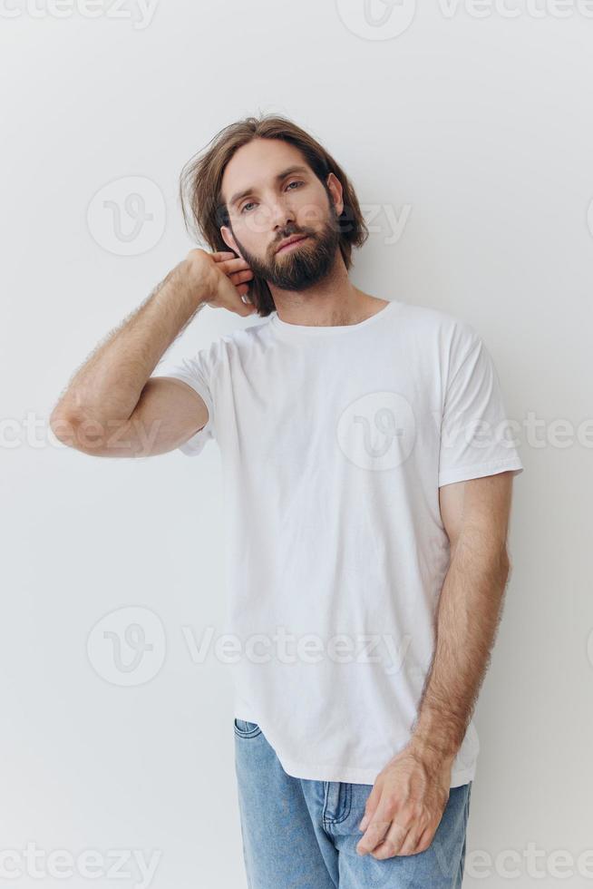 A man in a white T-shirt and blue jeans stands against a white wall and poses photo