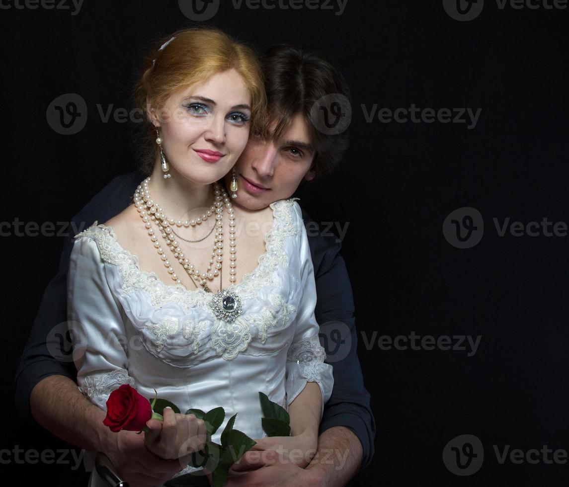 hermosa Pareja en retro estilo. un hombre abrazos un mujer. niña en un blanco vestir con un rojo Rosa en un antecedentes de un hombre. foto