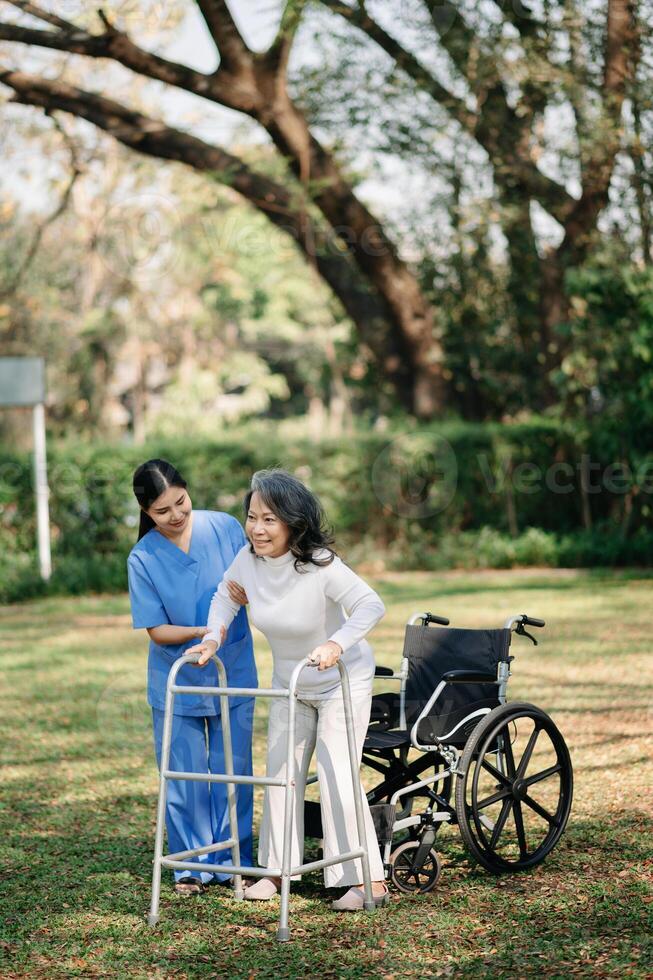 Asian careful caregiver or nurse and the happy patient in a wheelchair are walking in the garden to help and encourage and rest your mind with green nature. Help support yourself to learn to walk. photo