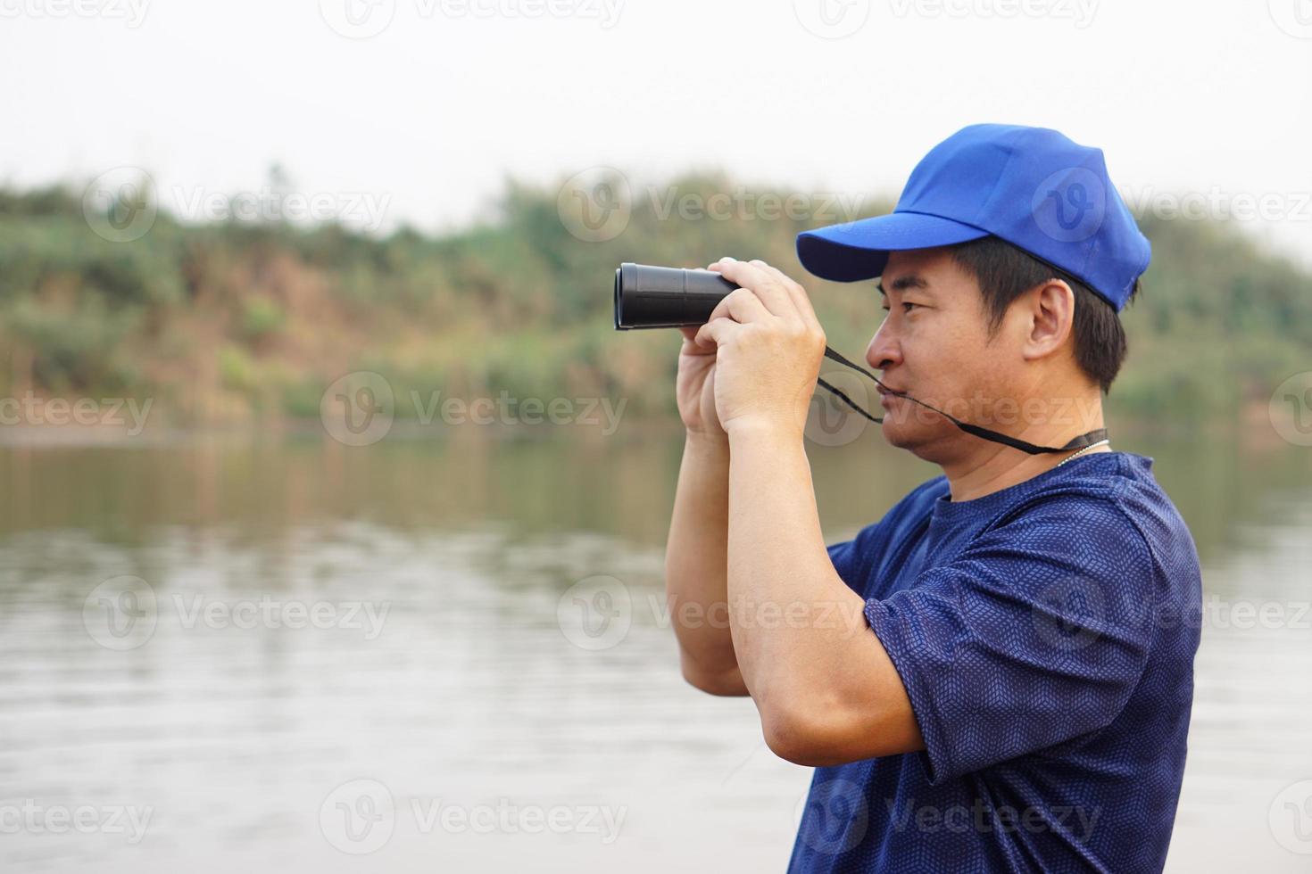 Asian man wears blue cap, hold binocular at the lake, nature source. Concept, nature exploration. Ecology study.  Pastime activity, lifestyle. Man explore environment photo