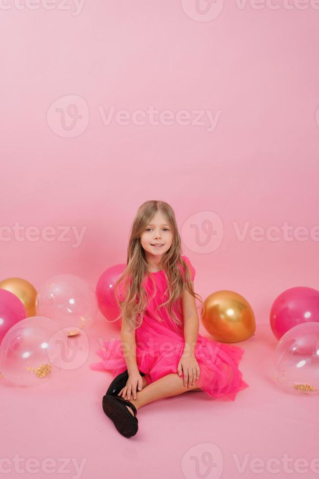 Smiling seven-year-old girl dressed in a fashionable dress sits on a pink background surrounded by balloons. Celebrate your birthday photo