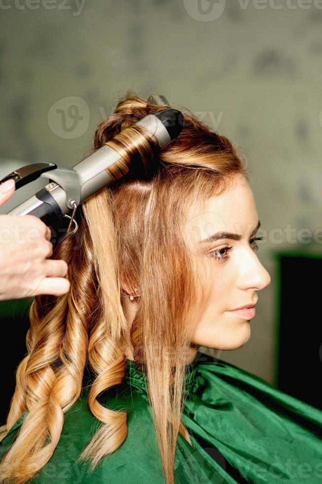 Hairdresser using curling tongs curls long brown hair on the young caucasian girl in a beauty salon. photo