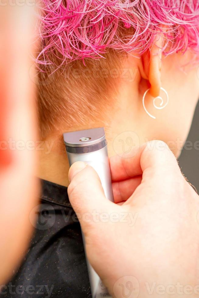 Back view of hairdresser's hand shaving nape and neck with electric trimmer of young caucasian woman with short pink hair in beauty salon. photo