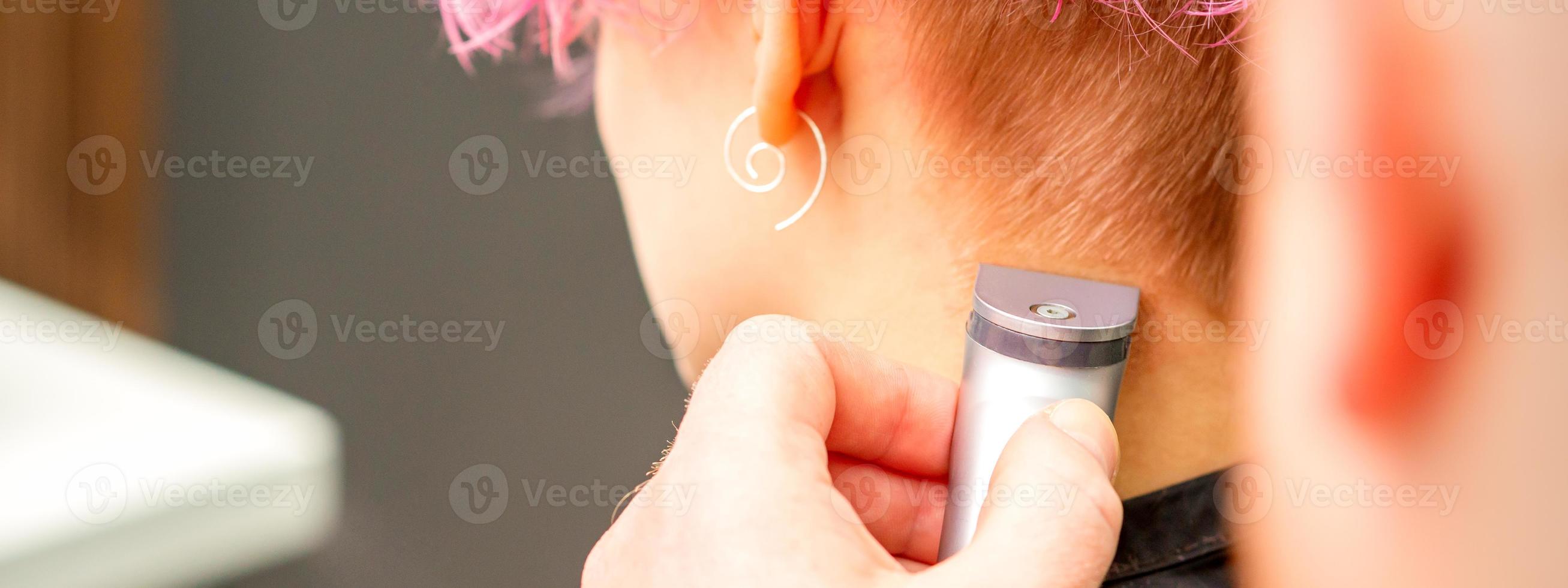 Back view of hairdresser's hand shaving nape and neck with electric trimmer of young caucasian woman with short pink hair in beauty salon. photo
