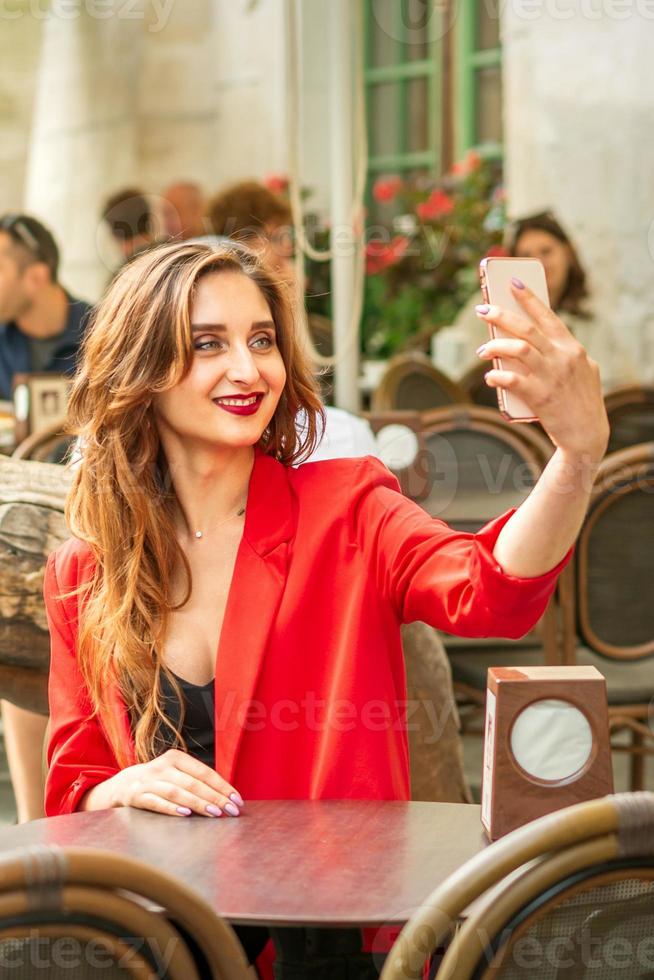 turista joven caucásico mujer en un rojo chaqueta con maleta toma un selfie a el mesa en café al aire libre. foto