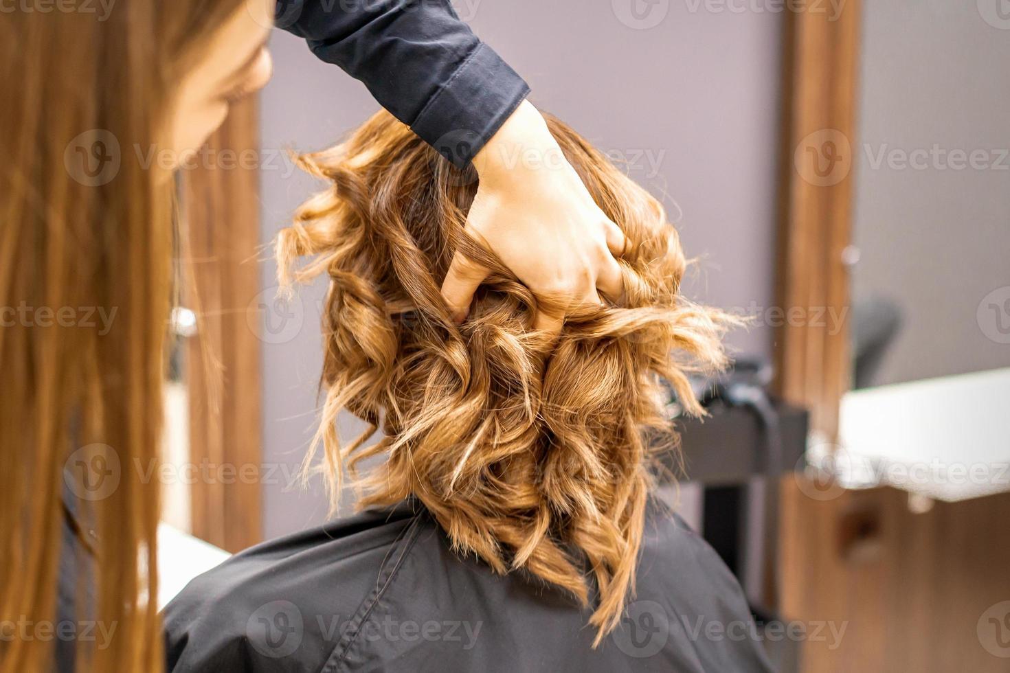 Female hairdresser checks brown curly hairstyle of a young caucasian woman in beauty salon. photo