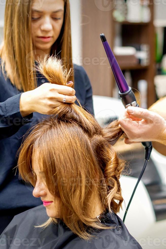 Two hairstylists using curling iron on customers long brown hair in a beauty salon. photo