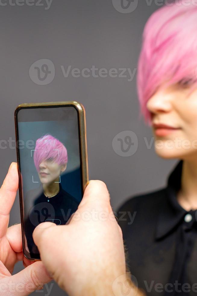 hombre peluquería manos tomando imagen en teléfono inteligente de su cliente corto rosado peinado. foto