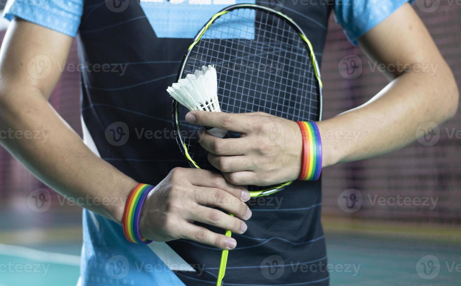 Badminton player wears rainbow wristbands and holding racket and white shuttlecock in front of the net before serving it to player in another side of the court, concept for LGBT people activities. photo