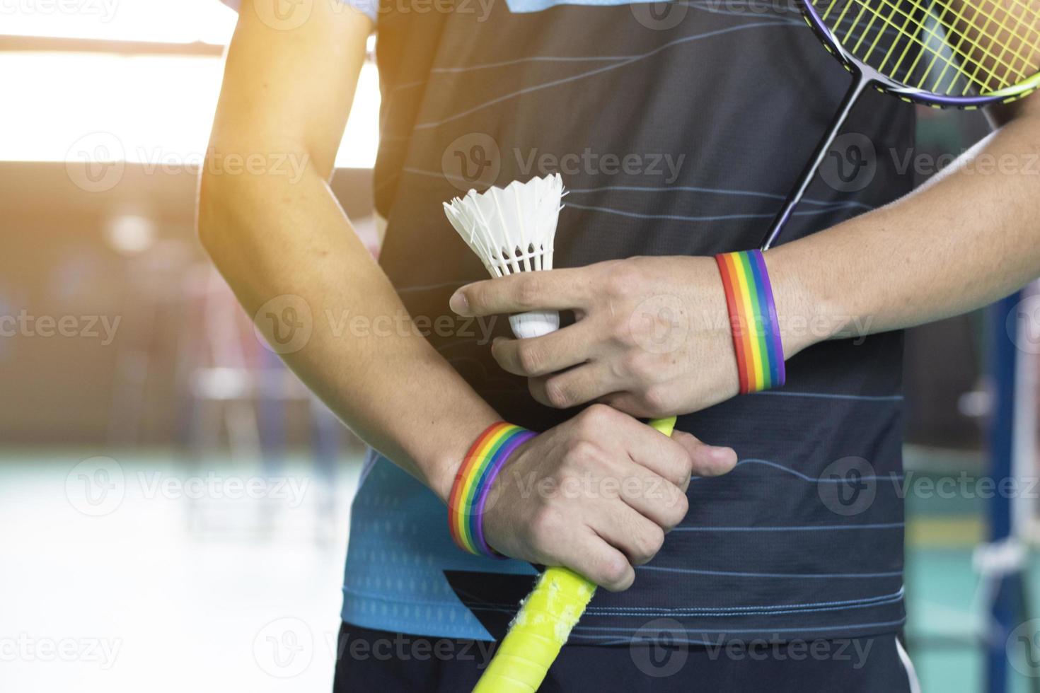 Badminton player wears rainbow wristbands and holding racket and white shuttlecock in front of the net before serving it to player in another side of the court, concept for LGBT people activities. photo