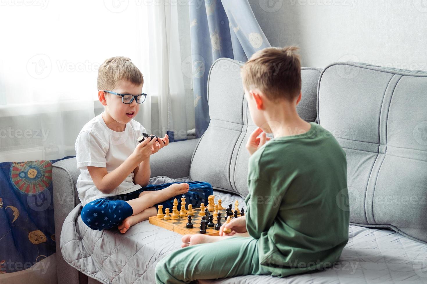 Boys playing wooden chess lying on a gray sofa in front of the window photo
