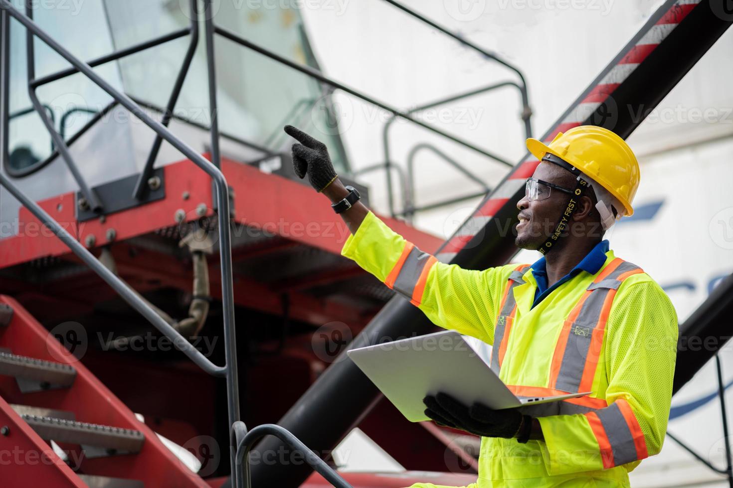 Portrait of African happy worker in protective safety jumpsuit uniform  with hardhat and use laptop computer at cargo container shipping warehouse. transportation import,export logistic industrial photo