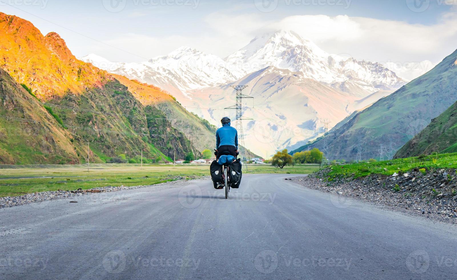 Male person in blue jacket cycling with touring bicycle surrounded by mountains and green summer nature. Tour around kazbegi national park. Georgia travel cycling holidays concept photo