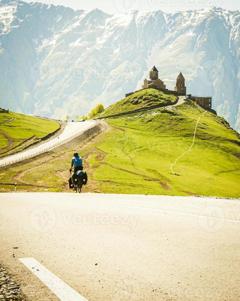 Male cyclist on touring bicycle cycling towards Gergeti trinity church with mountains background. Cycling holidays and travel around caucasus mountain range. photo