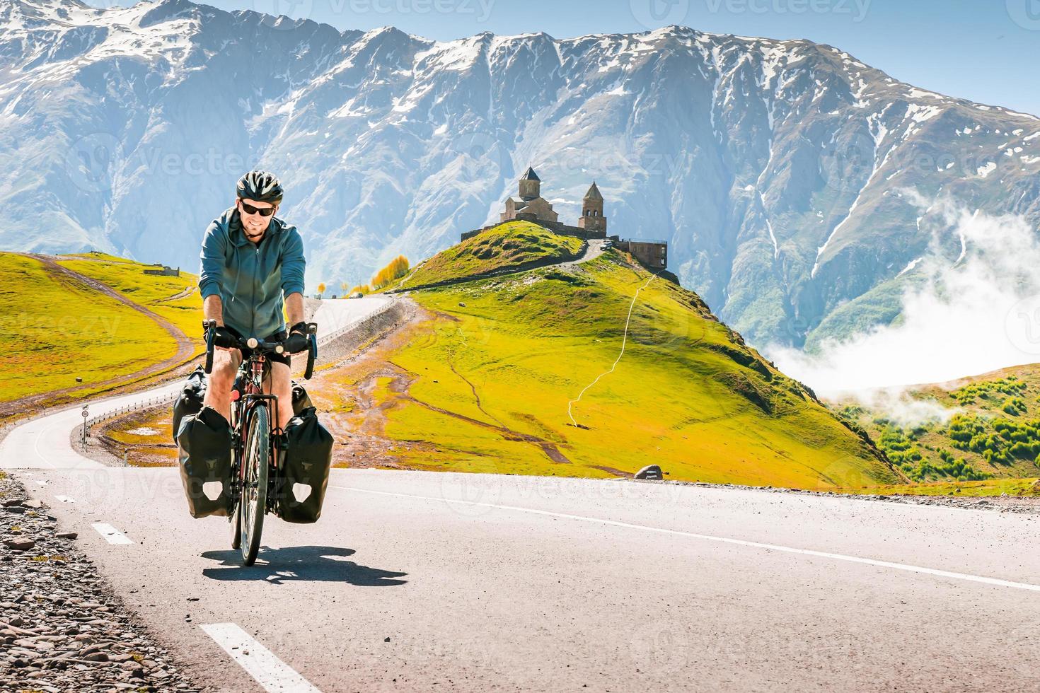 Cyclist back view on the road in scenic caucasus nature with Gergeti trinity monastery in the background. Traveller on bicycle. Solo travel long distance bicycle touring concept photo