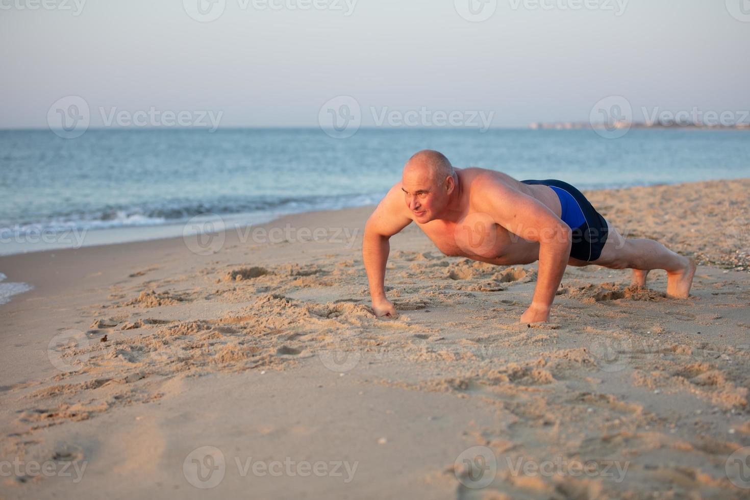 An elderly man is pushing up on the sea coast. Sports at sea middle-aged men. Healthy lifestyle on vacation. photo