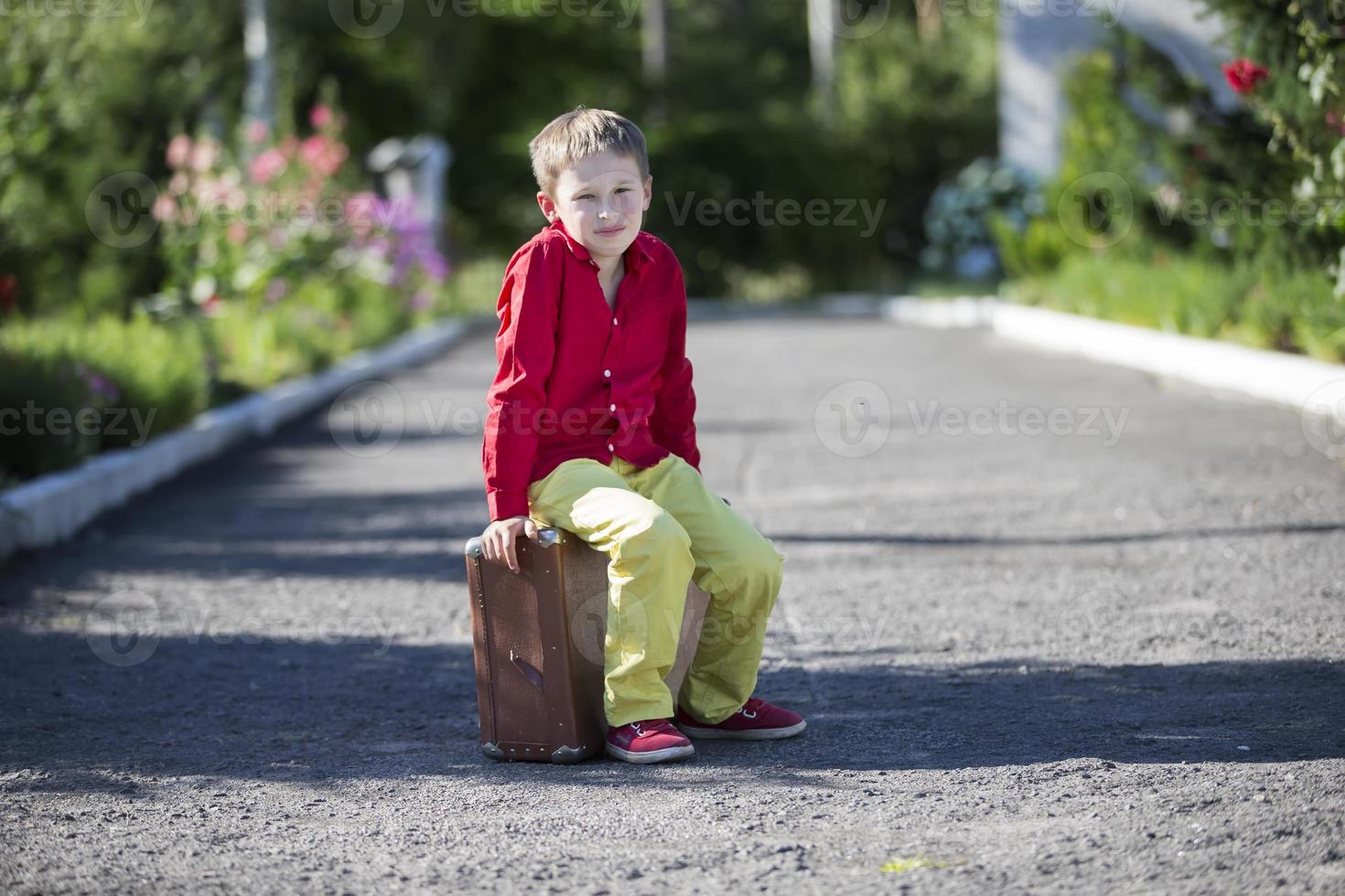 A sad boy sits on an old suitcase on the road. The child is about to leave. photo