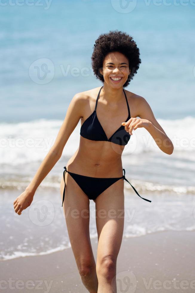 Joyful black woman in bikini walking along seaside photo