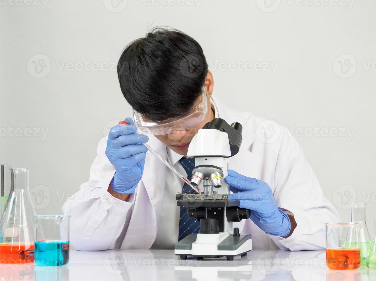 asiático masculino estudiante científico vistiendo un del doctor vestido en el laboratorio mirando mano a químico. causado por mezcla reactivos en científico investigación laboratorios con prueba tubos y microscopio en el mesa foto