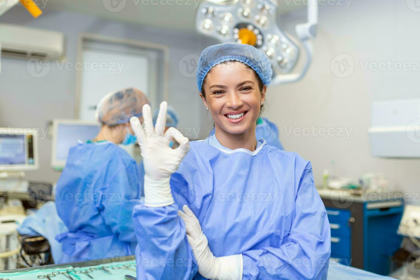 Portrait of female woman nurse surgeon OR staff member dressed in surgical scrubs gown mask and hair net in hospital operating room theater making eye contact smiling showing ok sign photo