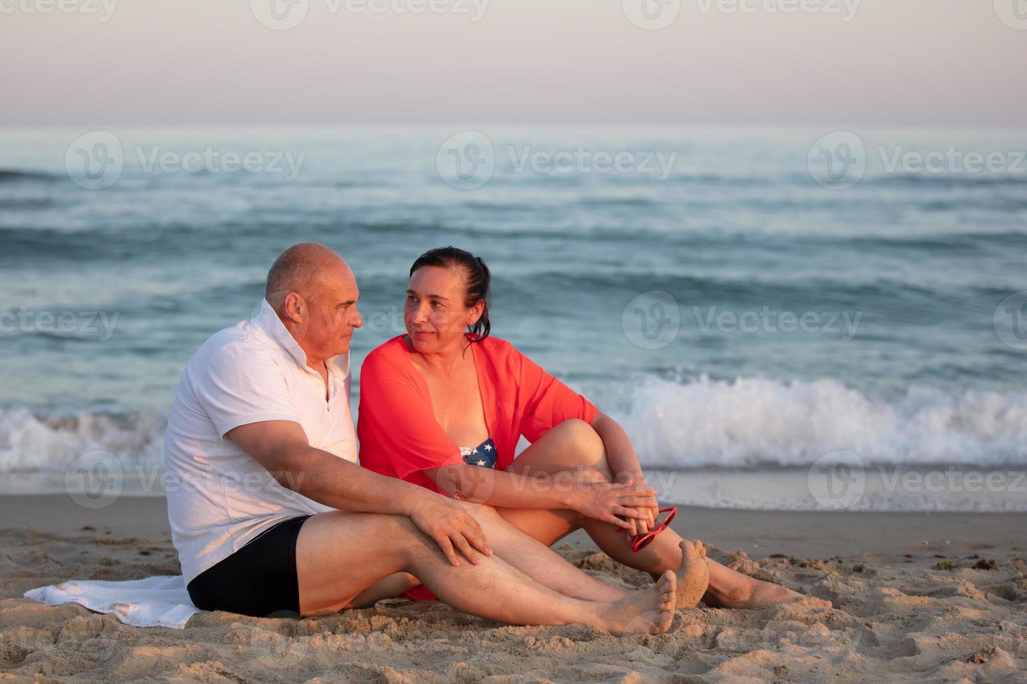 A middle-aged woman and man are sitting on a sandy shore against the background of the sea. Happy married couple at the resort. photo