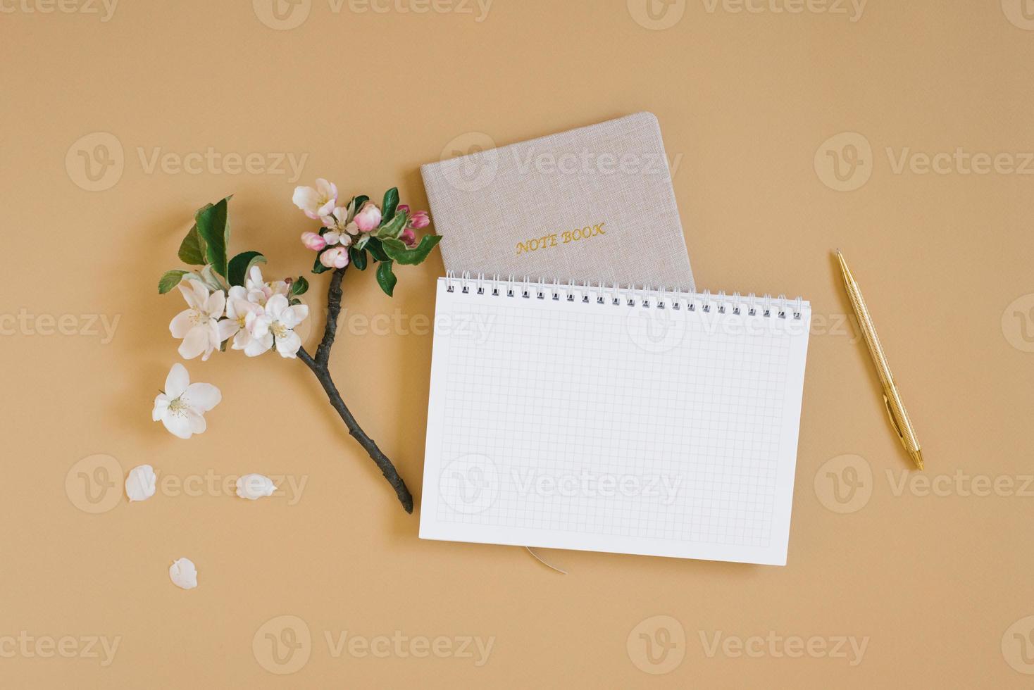 Open notebook with a blank white sheet of paper for copy space, notepad, pen and white flowers on a beige background. Flat lay workspace photo