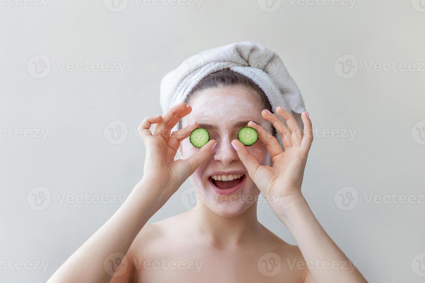 Beauty portrait of woman in towel on head with white nourishing mask or creme on face holding cucumber slices, white background isolated photo