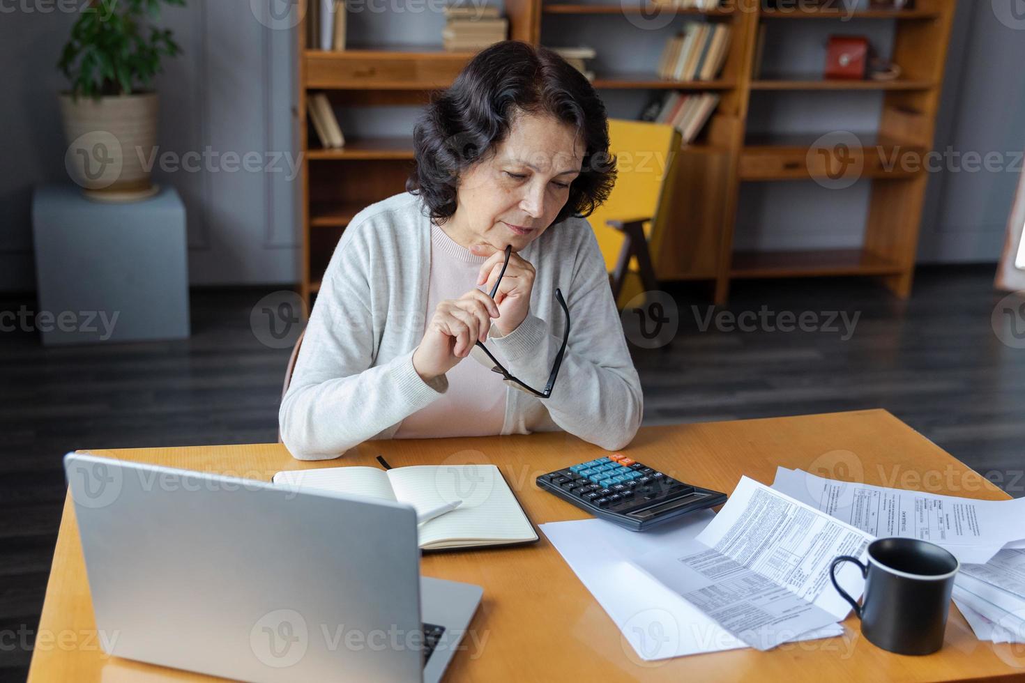 Middle aged senior woman sit with laptop and paper document, Pensive older mature lady reading paper bill pay online at home managing bank finances calculating taxes planning loan debt pension payment photo