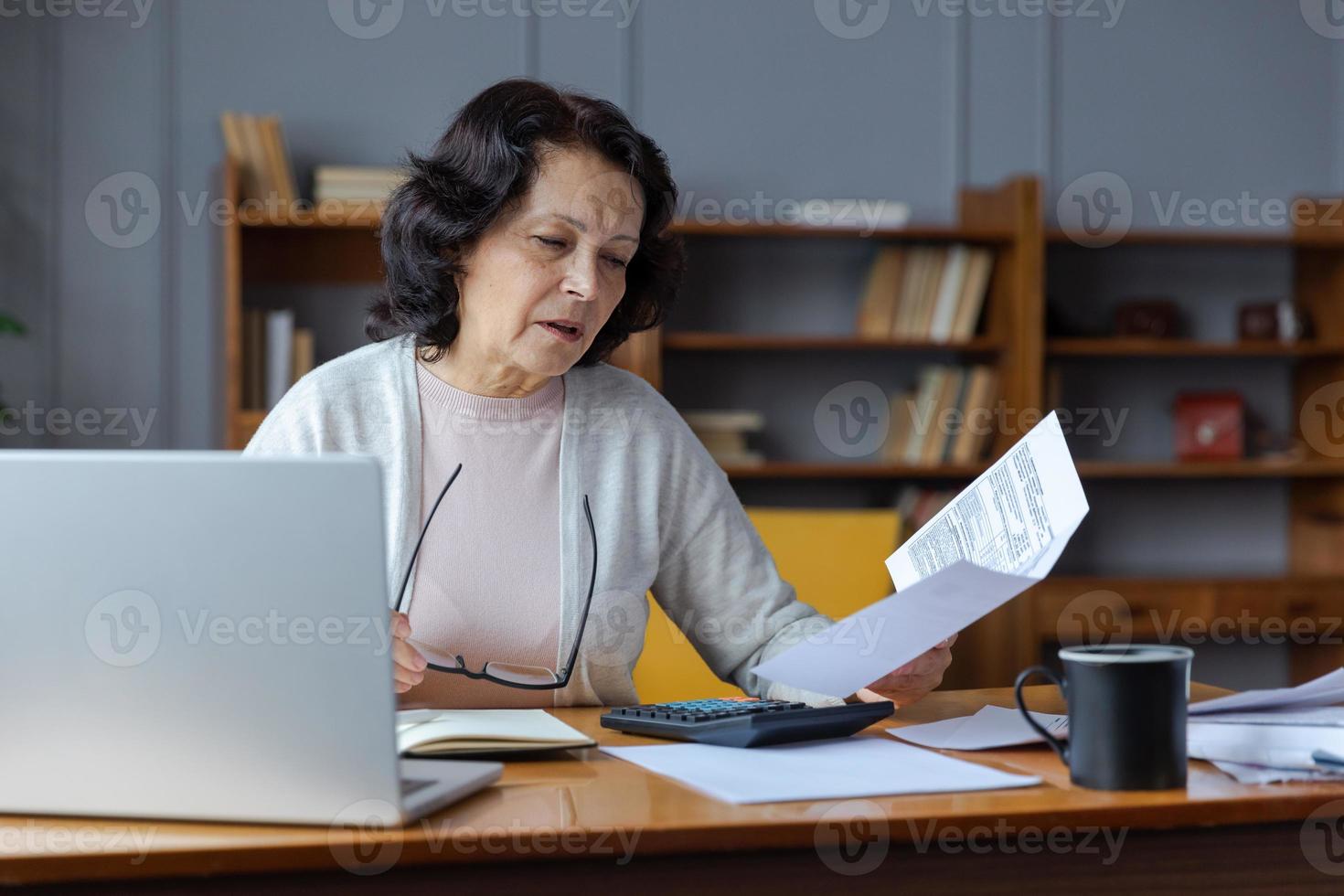 Middle aged senior woman sit with laptop and paper document, Pensive older mature lady reading paper bill pay online at home managing bank finances calculating taxes planning loan debt pension payment photo