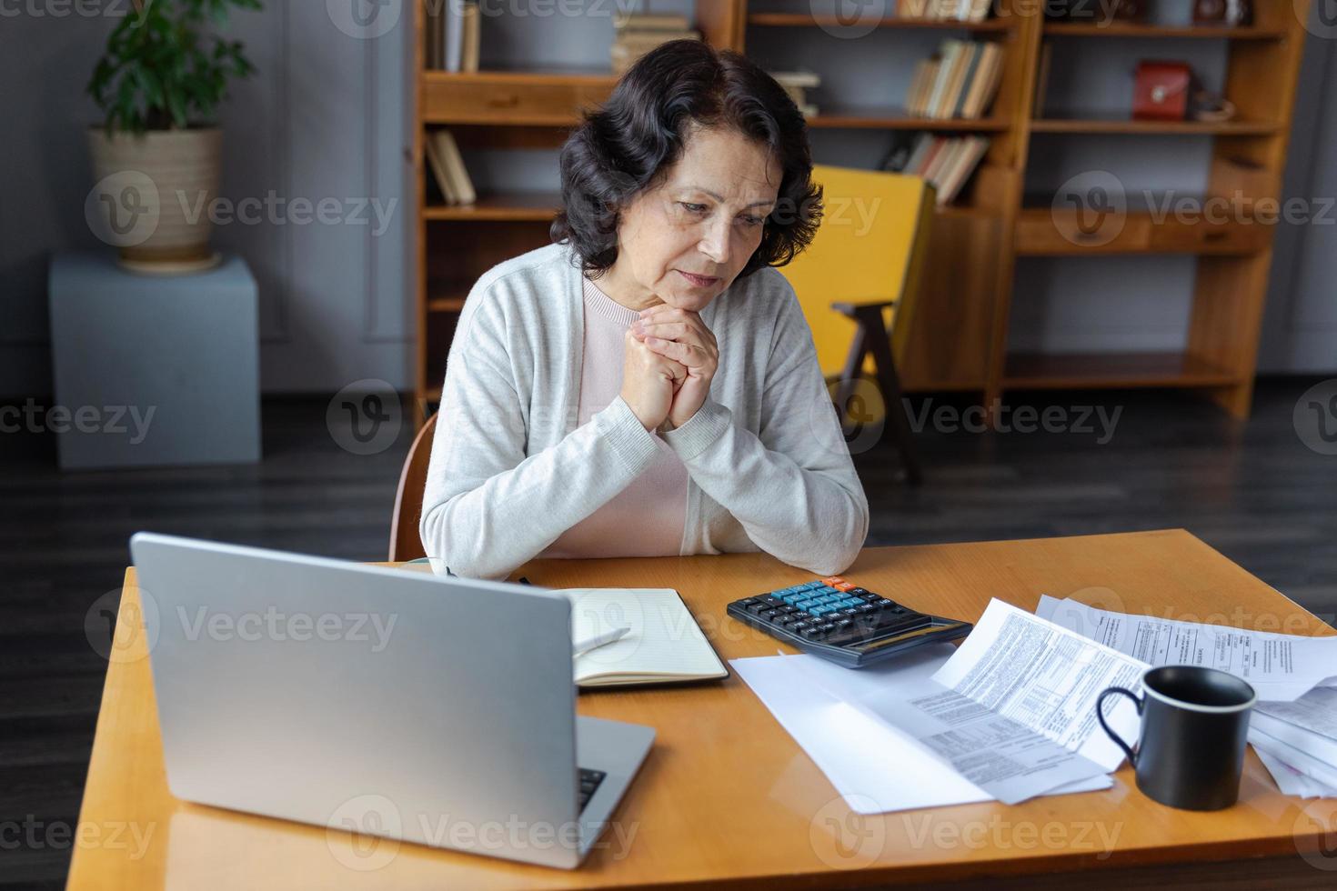 Middle aged senior woman sit with laptop and paper document, Pensive older mature lady reading paper bill pay online at home managing bank finances calculating taxes planning loan debt pension payment photo