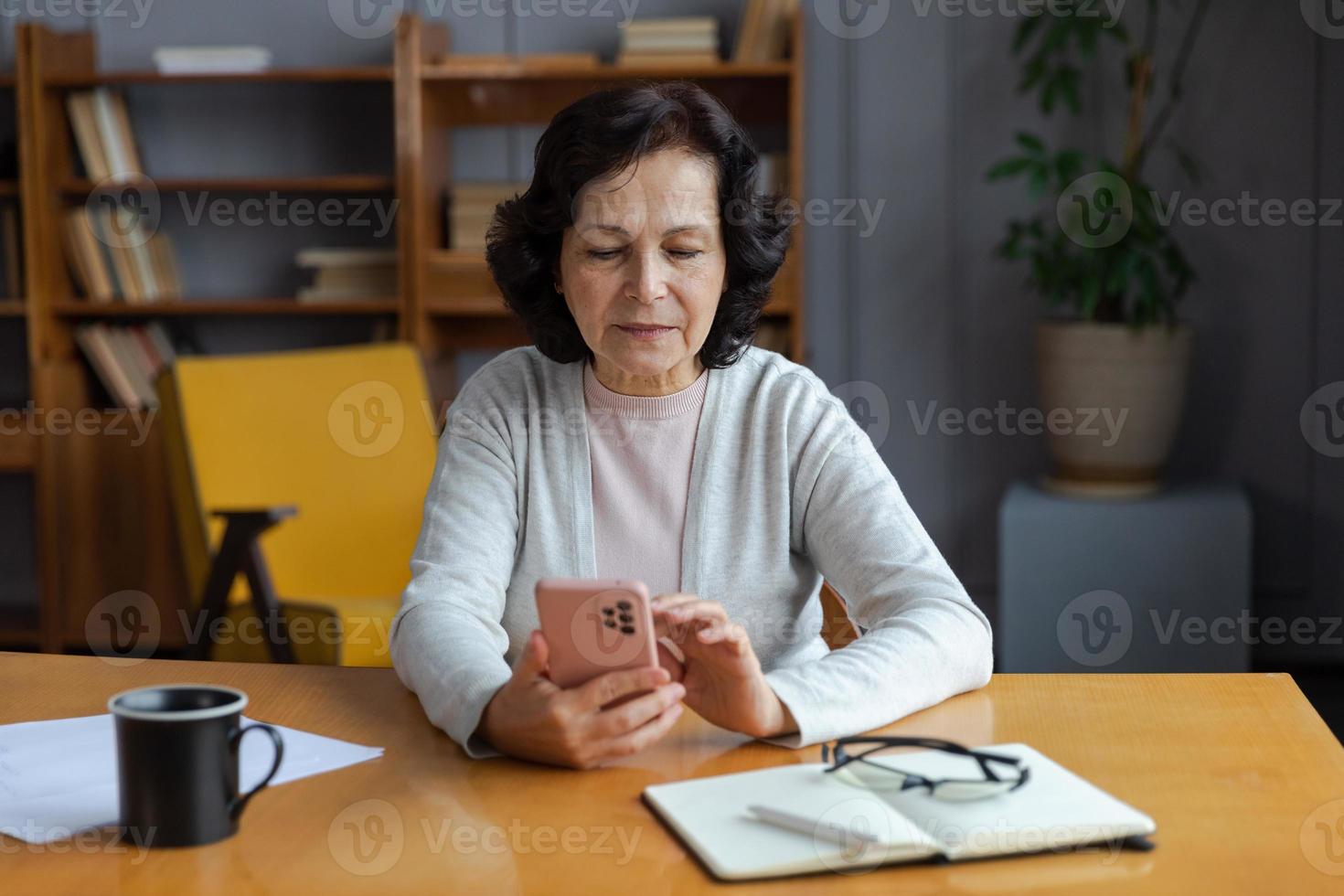 European middle aged senior woman holding using smartphone touch screen typing scroll page. Stylish older mature 60s lady with cell phone using internet social media apps at home. Shopping online. photo