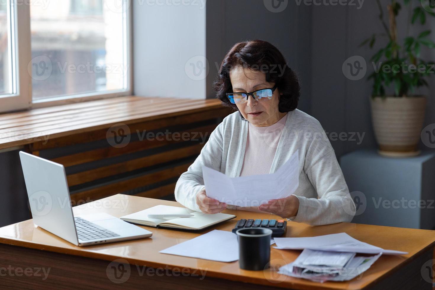 Middle aged senior woman sit with laptop and paper document, Pensive older mature lady reading paper bill pay online at home managing bank finances calculating taxes planning loan debt pension payment photo
