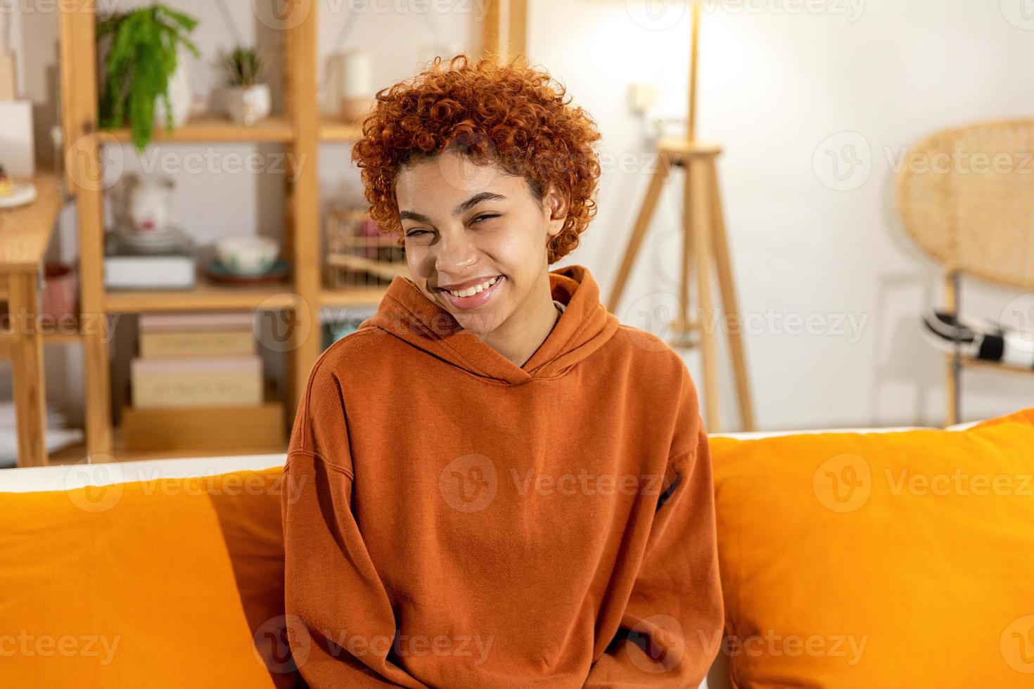 Beautiful african american girl with afro hairstyle smiling sitting on sofa at home indoor. Young african woman with curly hair laughing. Freedom happiness carefree happy people concept. photo