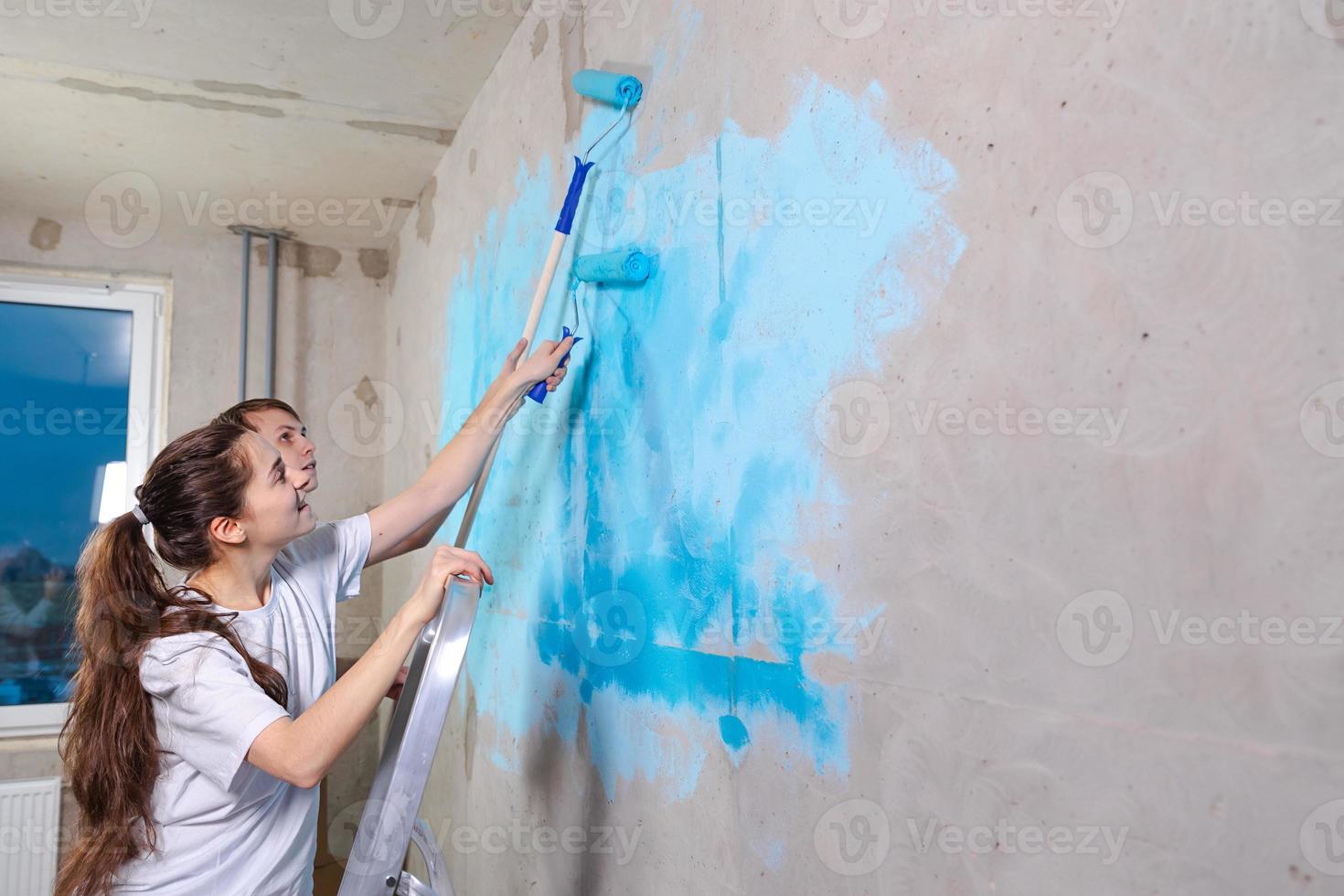 Couple in new home during repair works painting wall together. Happy family holding paint roller painting wall with blue color paint in new house. Home renovation DIY renew home concept. photo