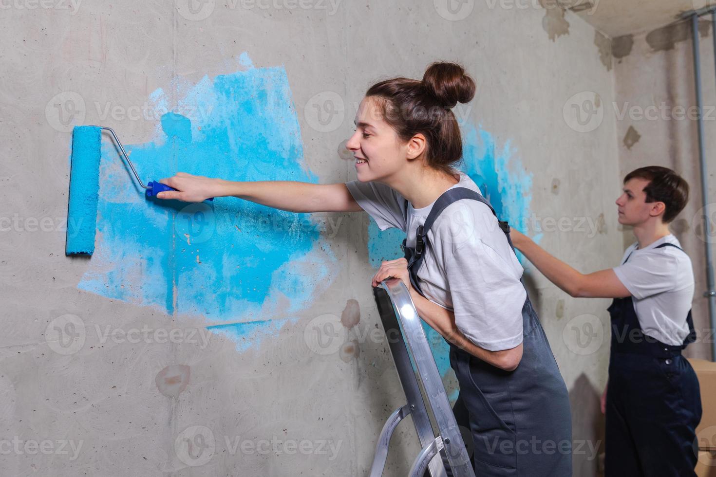 Couple in new home during repair works painting wall together. Happy family holding paint roller painting wall with blue color paint in new house. Home renovation DIY renew home concept. photo