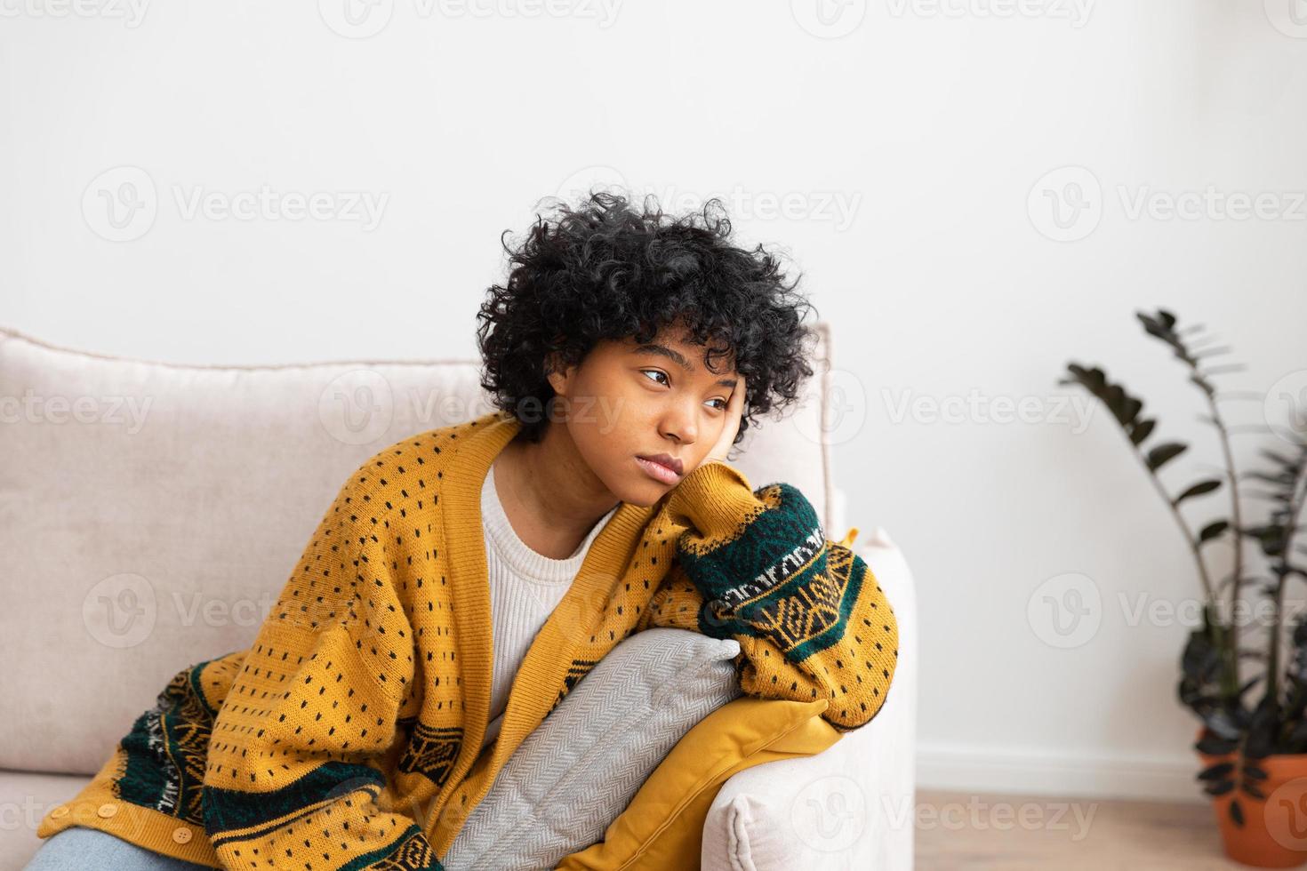 African american sad thoughtful pensive unmotivated girl sitting on sofa at home indoor. Young african woman ponder look tired after long day. Girl feels depressed offended lonely upset heartbreak. photo