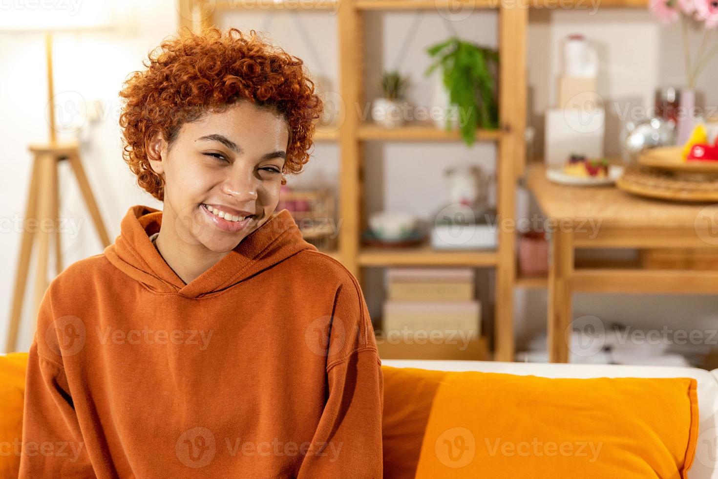 Beautiful african american girl with afro hairstyle smiling sitting on sofa at home indoor. Young african woman with curly hair laughing. Freedom happiness carefree happy people concept. photo