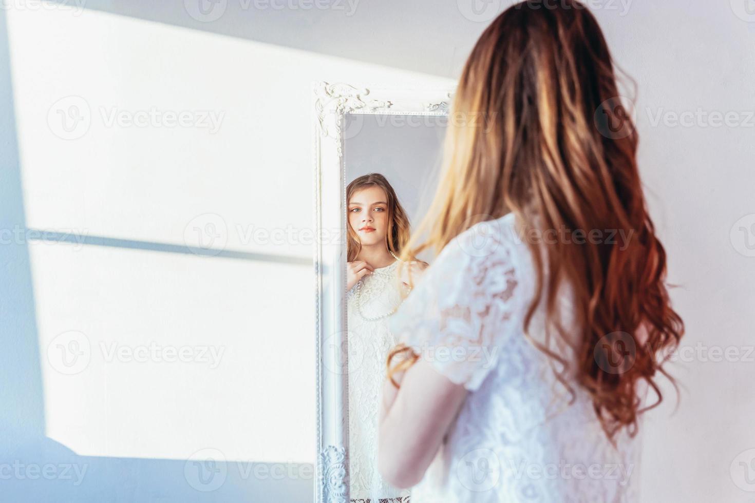 belleza maquillaje Mañana rutina amor tú mismo concepto. joven Adolescente niña mirando a reflexión en espejo. joven positivo mujer vistiendo blanco vestir posando en brillante ligero habitación en contra blanco pared. foto