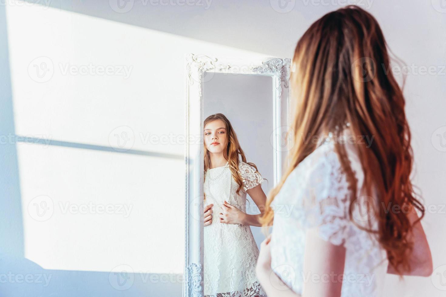 Beauty makeup morning rutine love yourself concept. Young teenage girl looking at reflection in mirror. Young positive woman wearing white dress posing in bright light room against white wall. photo