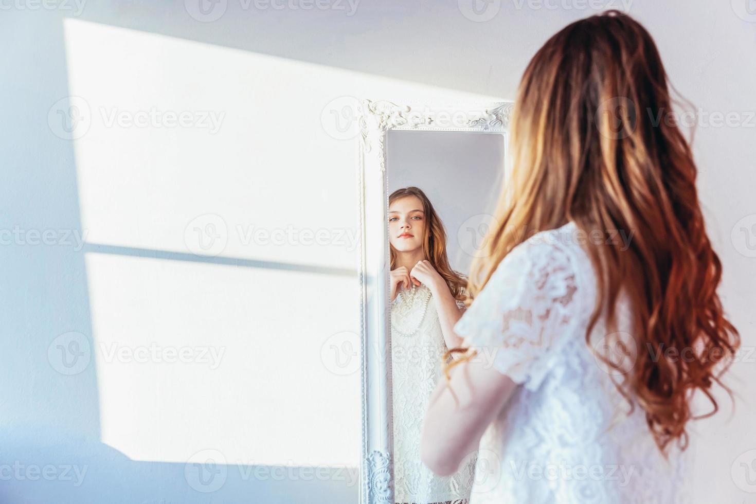 belleza maquillaje Mañana rutina amor tú mismo concepto. joven Adolescente niña mirando a reflexión en espejo. joven positivo mujer vistiendo blanco vestir posando en brillante ligero habitación en contra blanco pared. foto