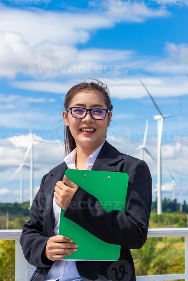 energía ingeniero hembra trabajando a viento granja. asiático negocio mujer vistiendo traje participación tableta y portapapeles con molino ambiente renovable energía. foto