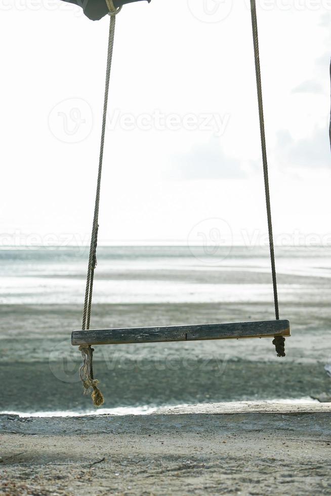 Selective focus on the old wooden swing hanging from the branch of tree isolated on the bright sky and seascape in background photo