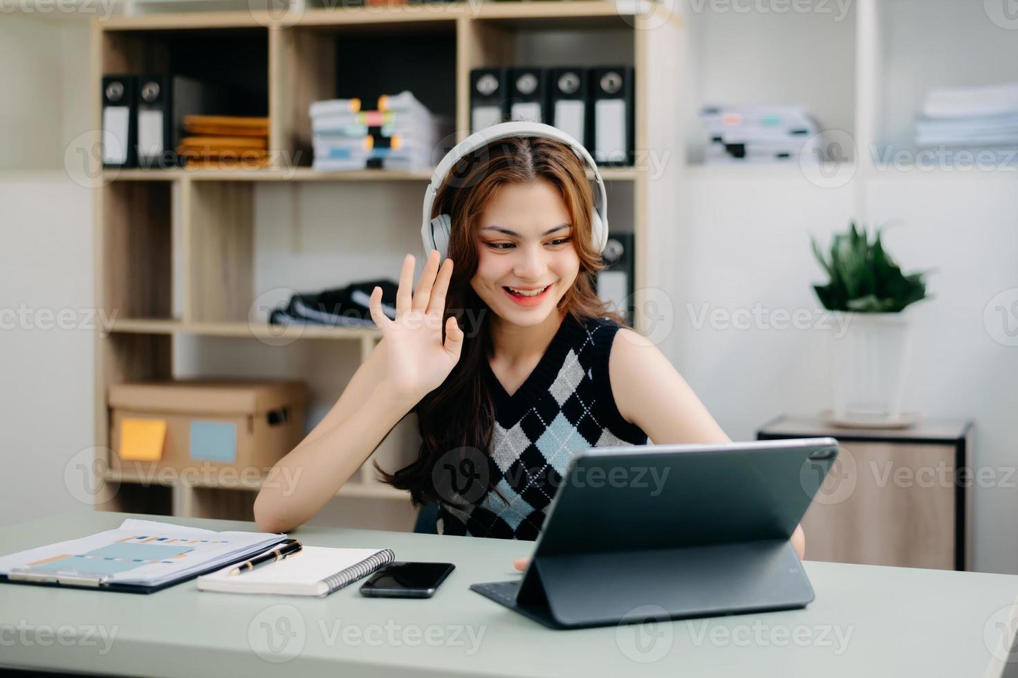 Attractive happy young student studying at the college library, sitting at the desk, using a laptop computer, tablet and headphones having a video chat. photo