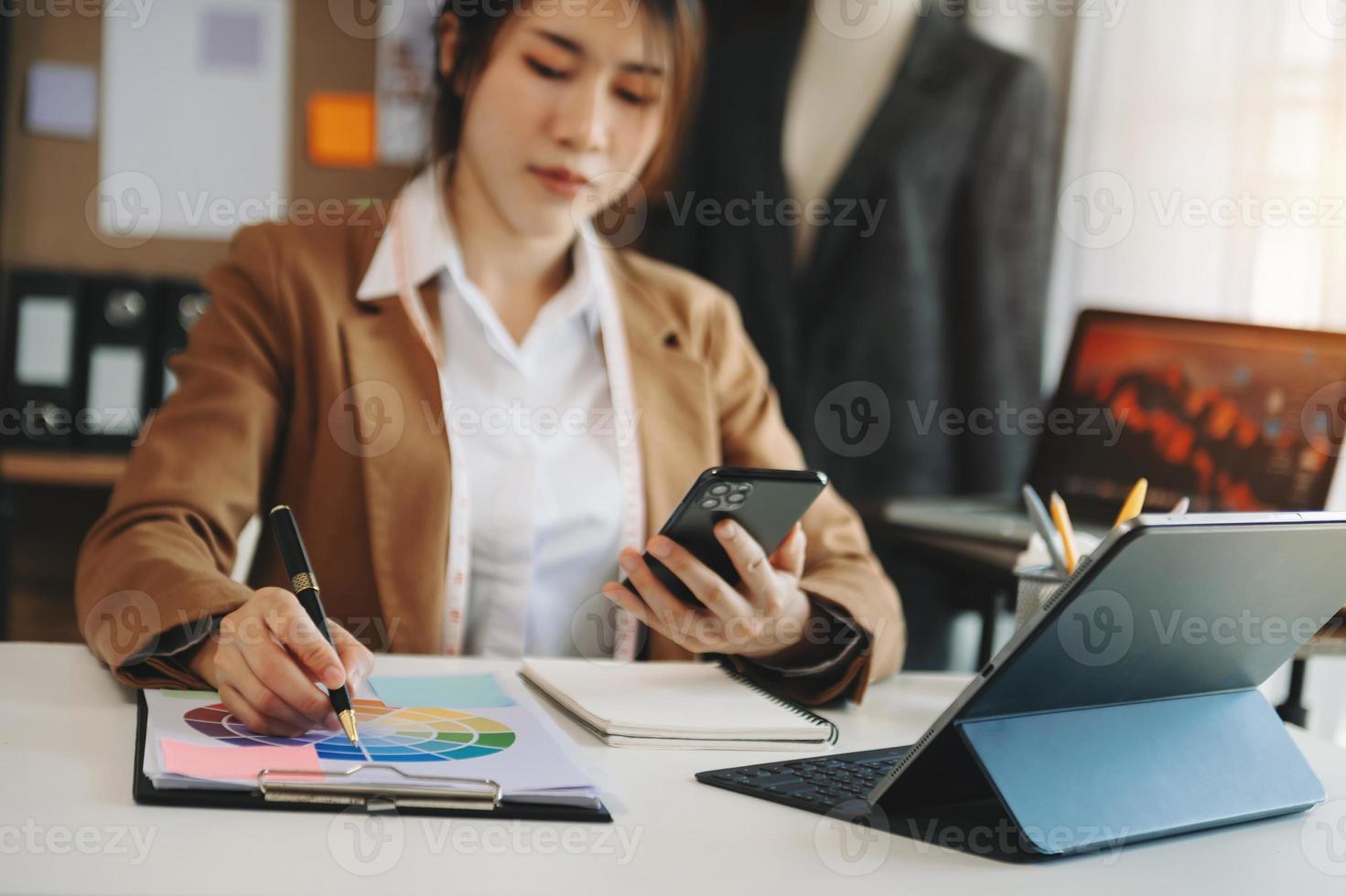 Moda diseñador l joven asiático mujer trabajando utilizando computadora portátil, tableta y sonriente mientras en pie en taller respondiendo foto