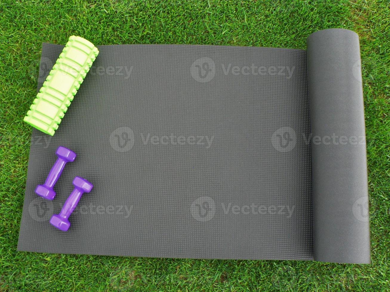 Keeping fit and exercising outdoor or at home. Purple dumbbells, green foam roll on a yoga mat on green grass lawn in a backyard or park. Healthy lifestyle. Copy space. photo