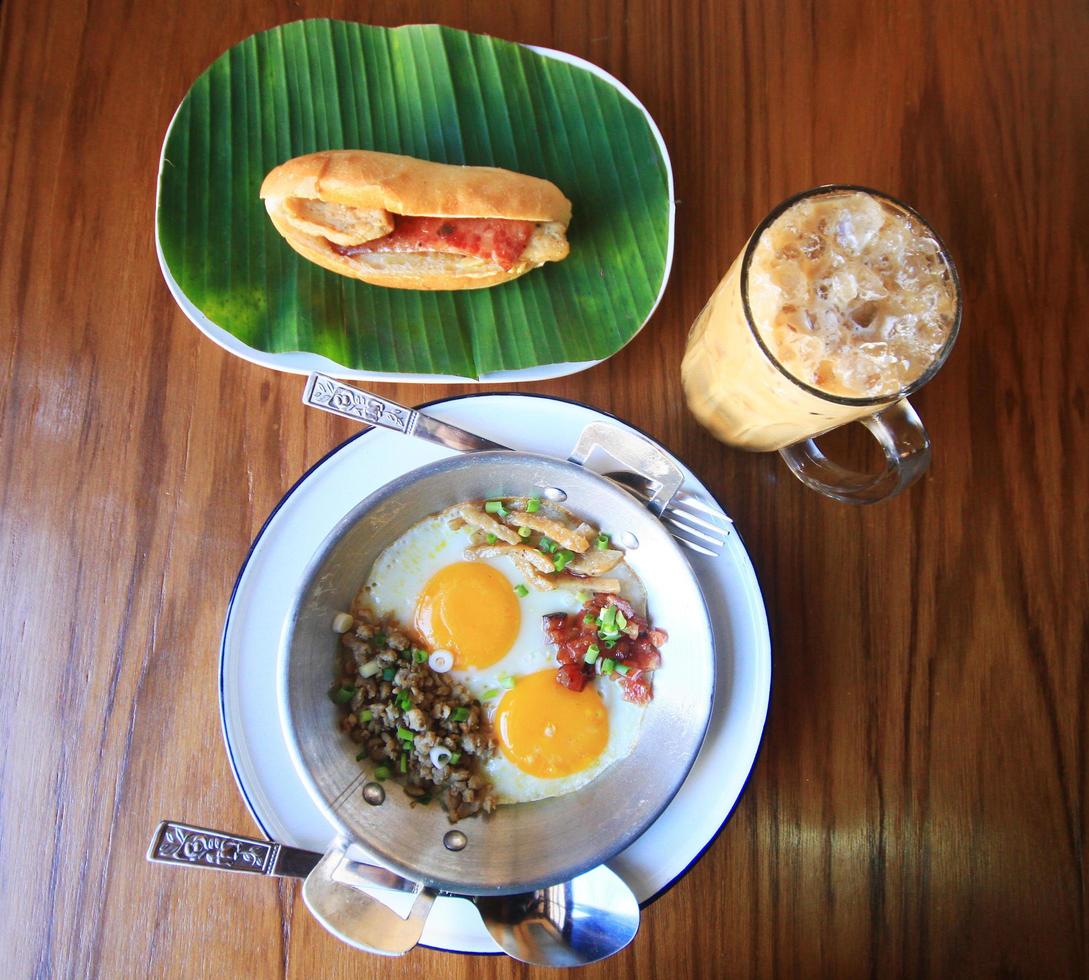 Indochina pan-fried egg with toppings with Baguette bread sandwich with cheese, ham on fresh Green banana leaf and ice coffee on wooden table in homemade Thai style photo