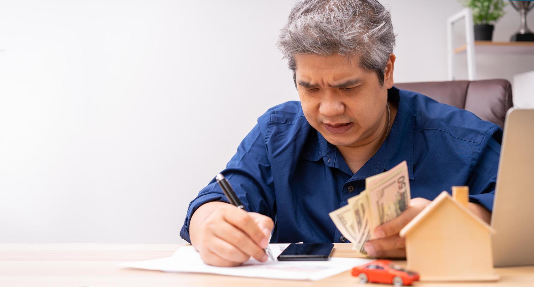 Unhappy Asian man holding a banknote and Making an account of income and expenses to pay the home loan and car loan, Concept loan payment and home insurance, economic problems, and inflation photo