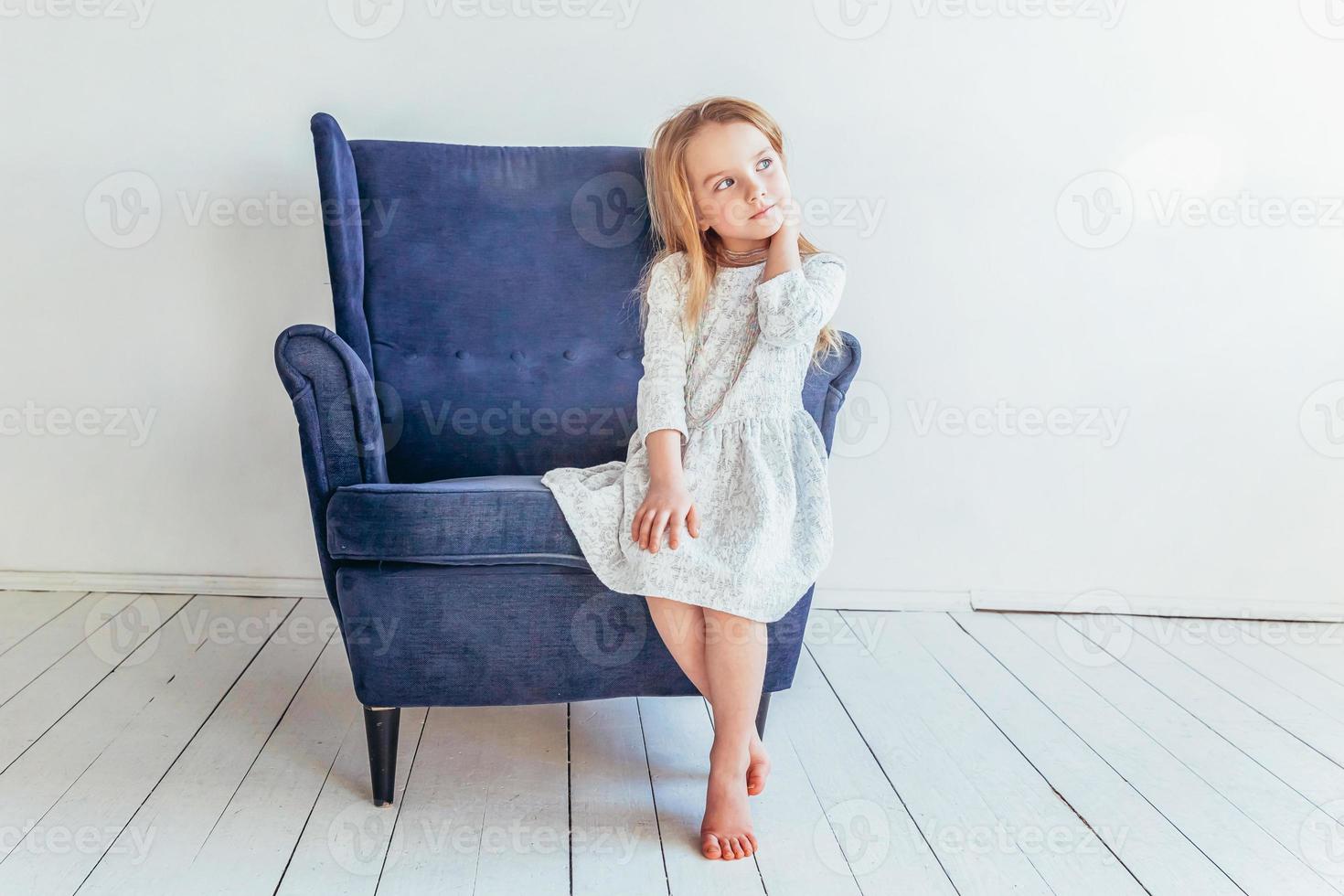 dulce pequeño niña en blanco vestir sentado en moderno acogedor azul silla relajante en blanco brillante vivo habitación a hogar adentro. infancia Niños de escuela juventud relajarse concepto. foto